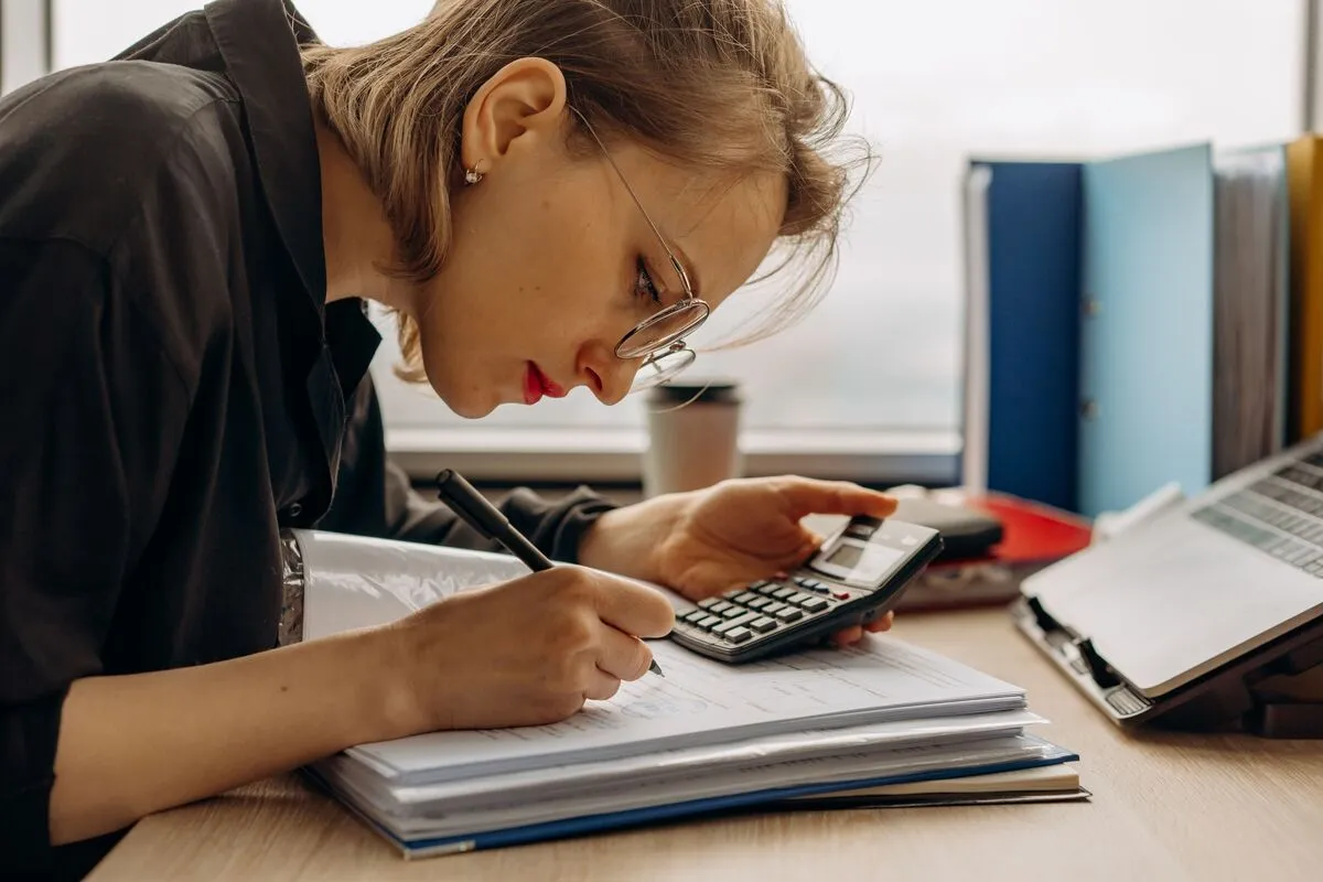 A woman in glasses doing the books with a calculator, a ledger, and folders.