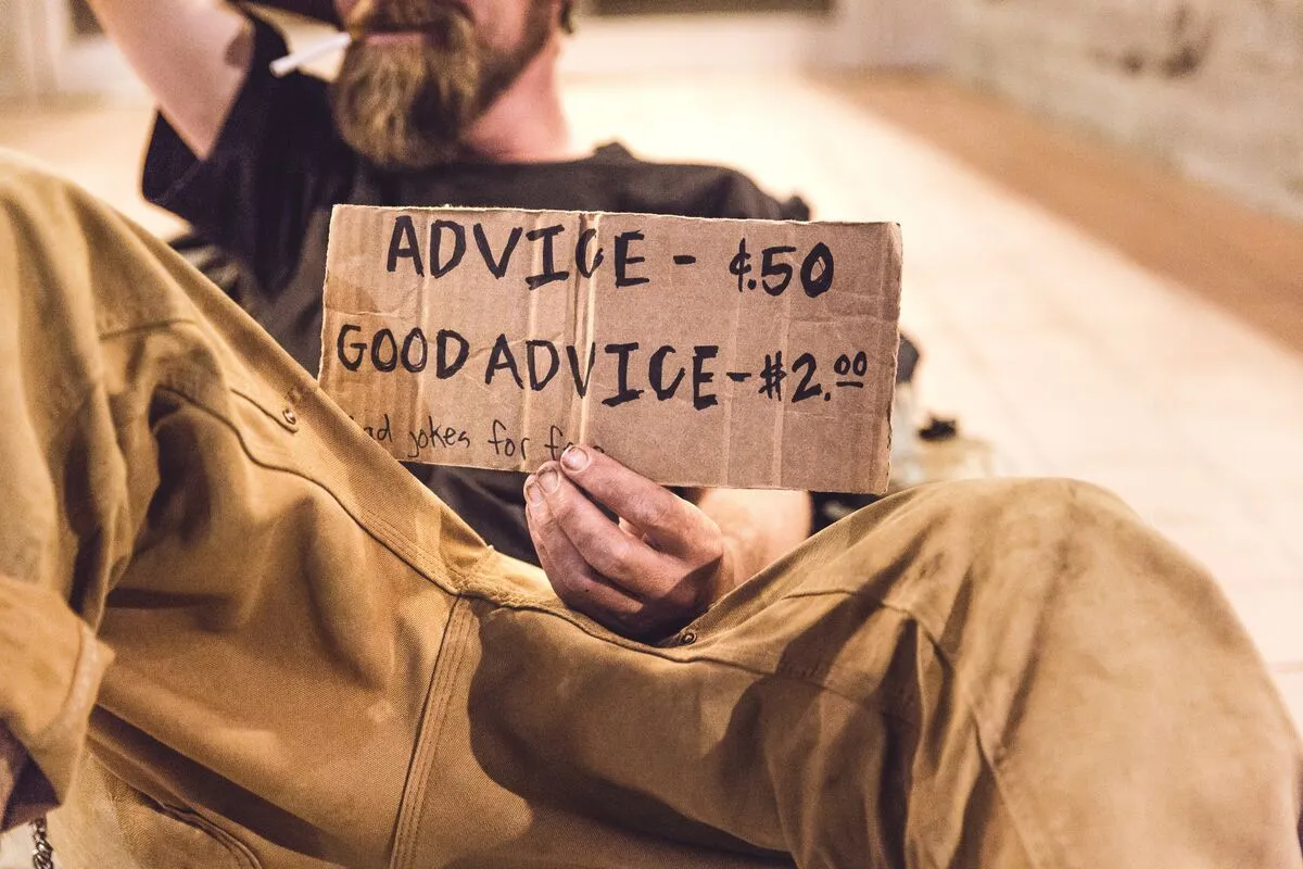A man in work overalls holding a cardboard sign reading "Advice $.50, Good advice $2.00".
