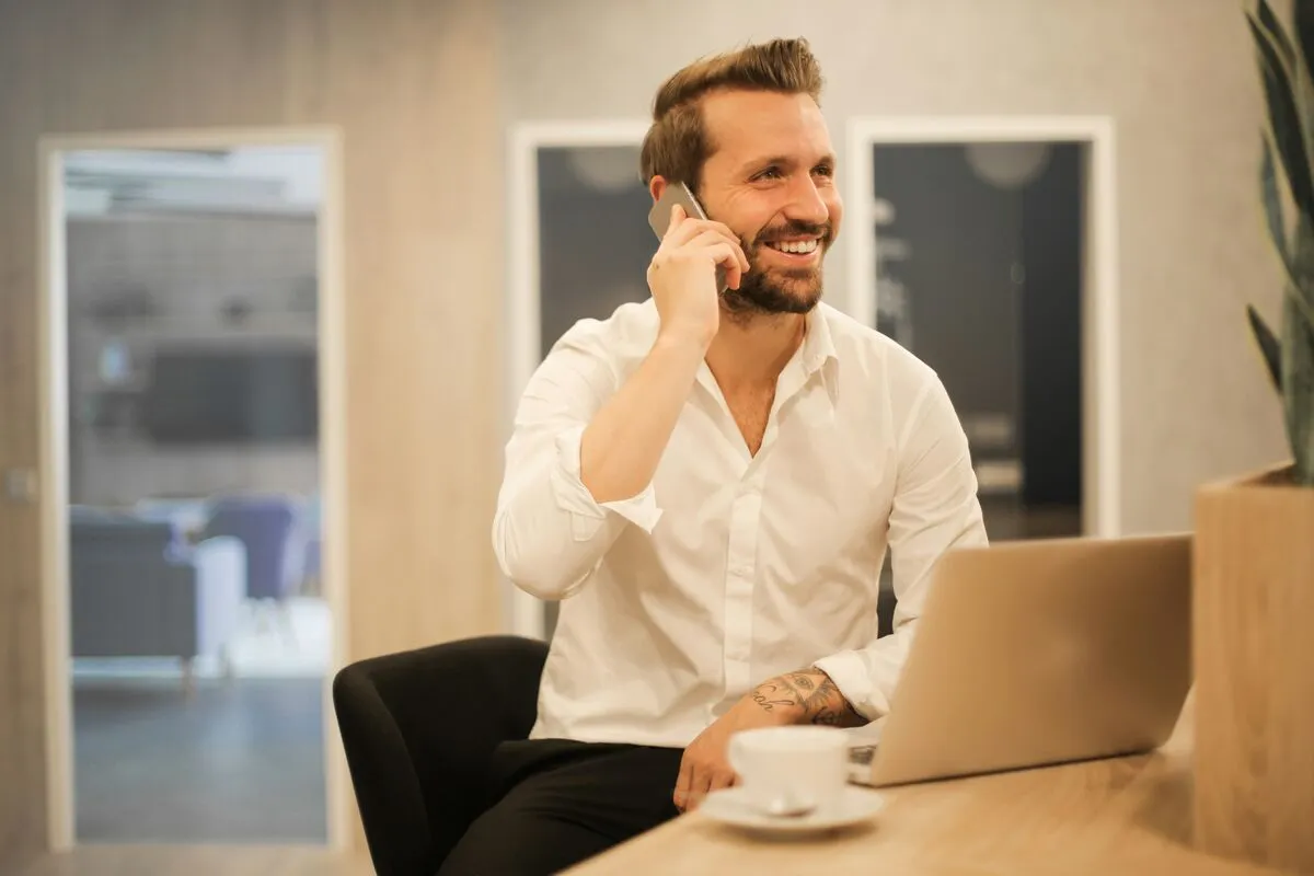 A smiling man in a white shirt takes a phone call at his laptop, a coffee on the desk in front of him.