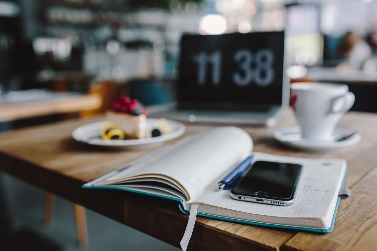 An open notebook and phone on a cafe table, with a laptop, coffee and cake behind.