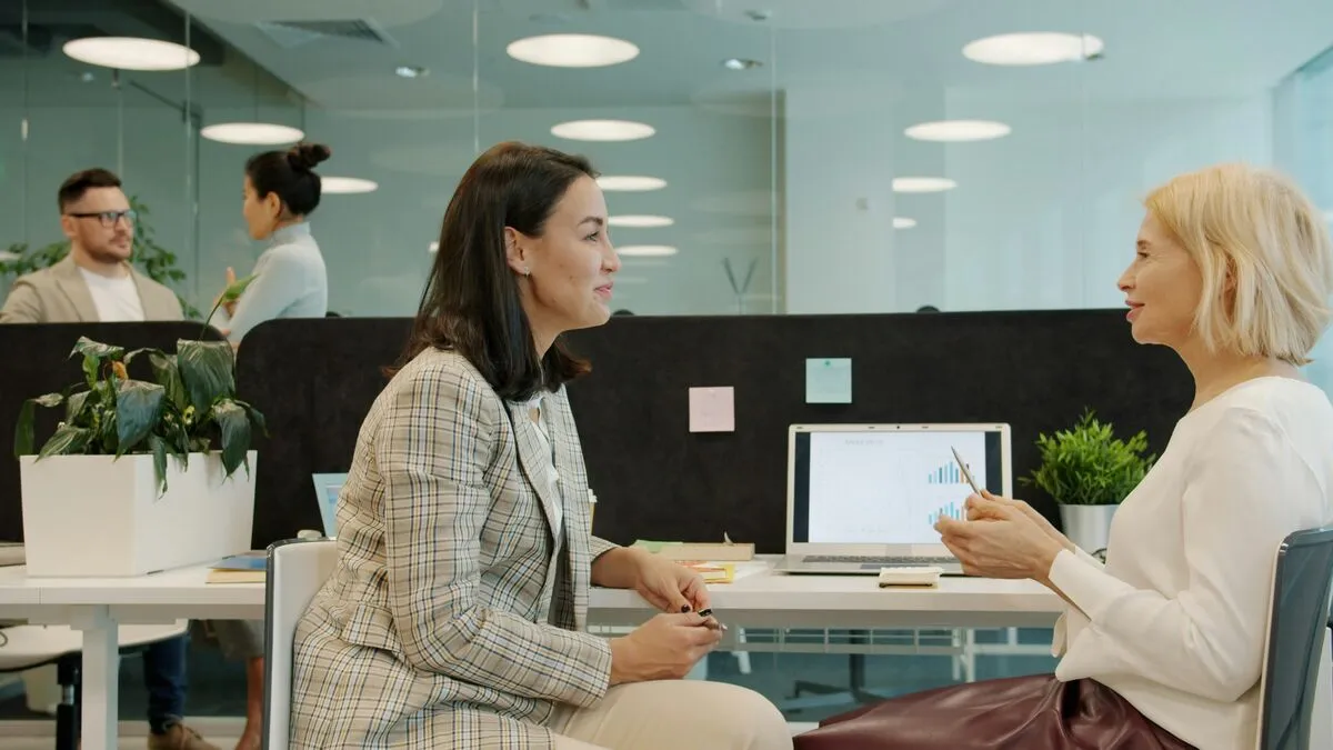 Two people in a consultation across a desk, one explaining while the other takes notes on a laptop.