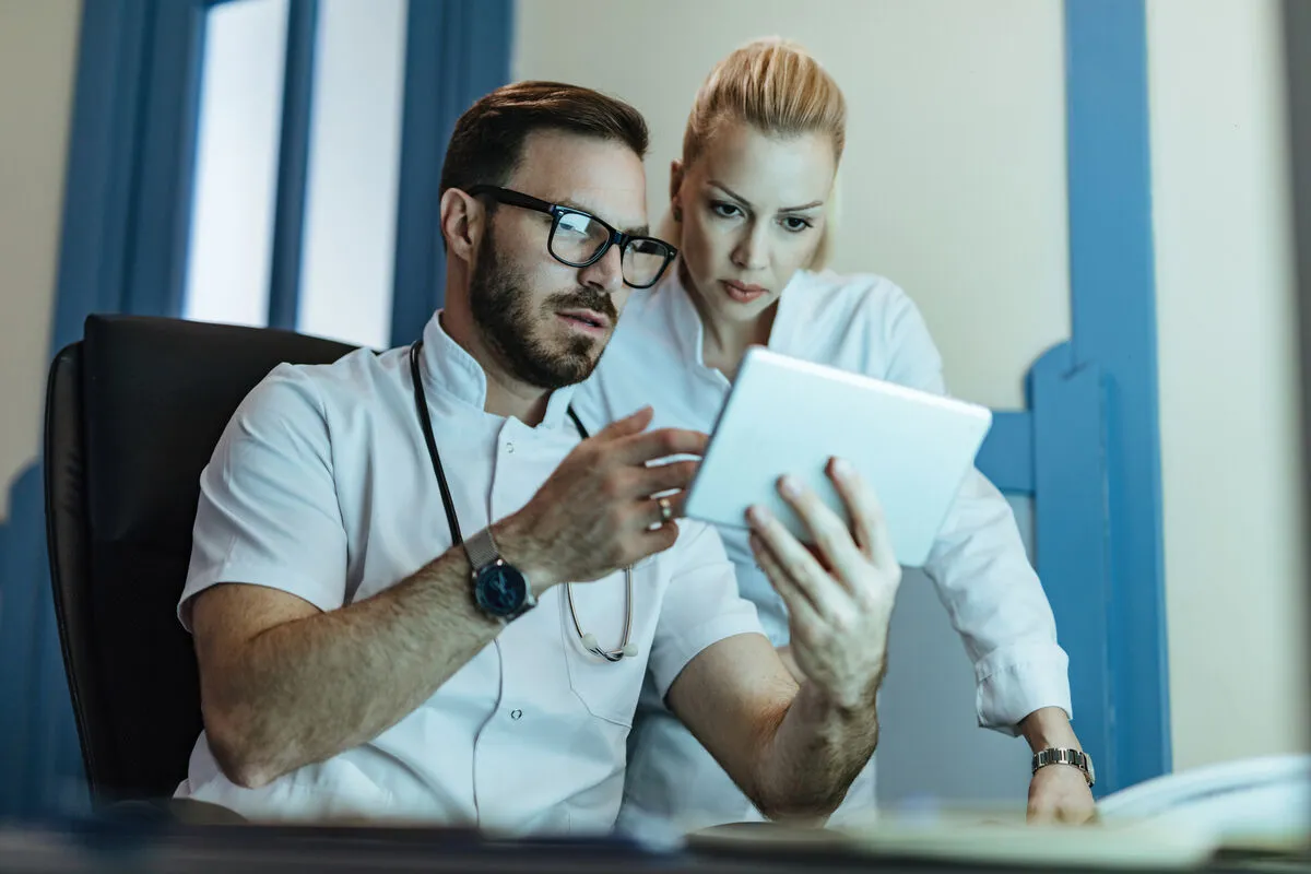 Two clinicians in white coats study a tablet together in a bright clinic.