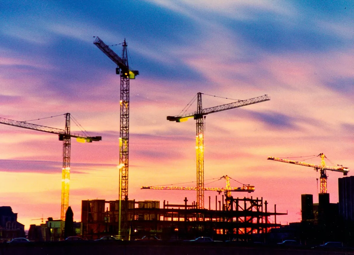 Construction cranes silhouetted over a building site against a sunset sky.
