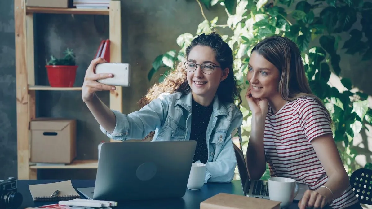 Two women at a desk take a photo on a phone, with a laptop, notebooks and pot plants around them.