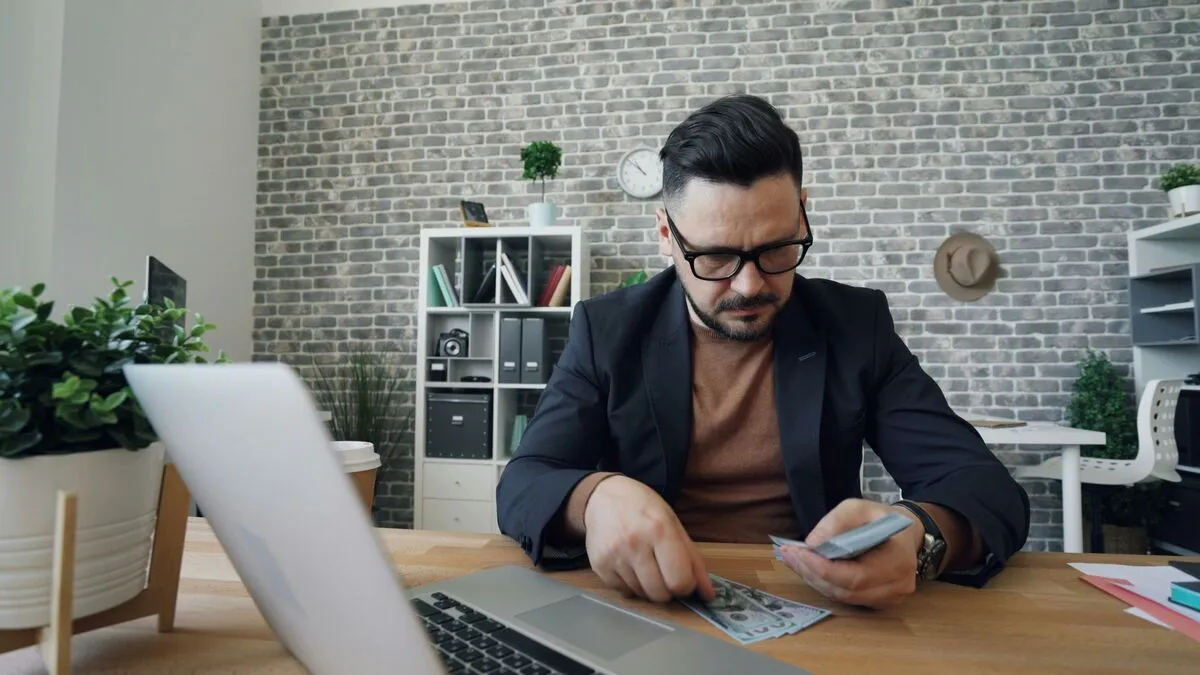 A man in a blazer counting cash at his desk beside a laptop.
