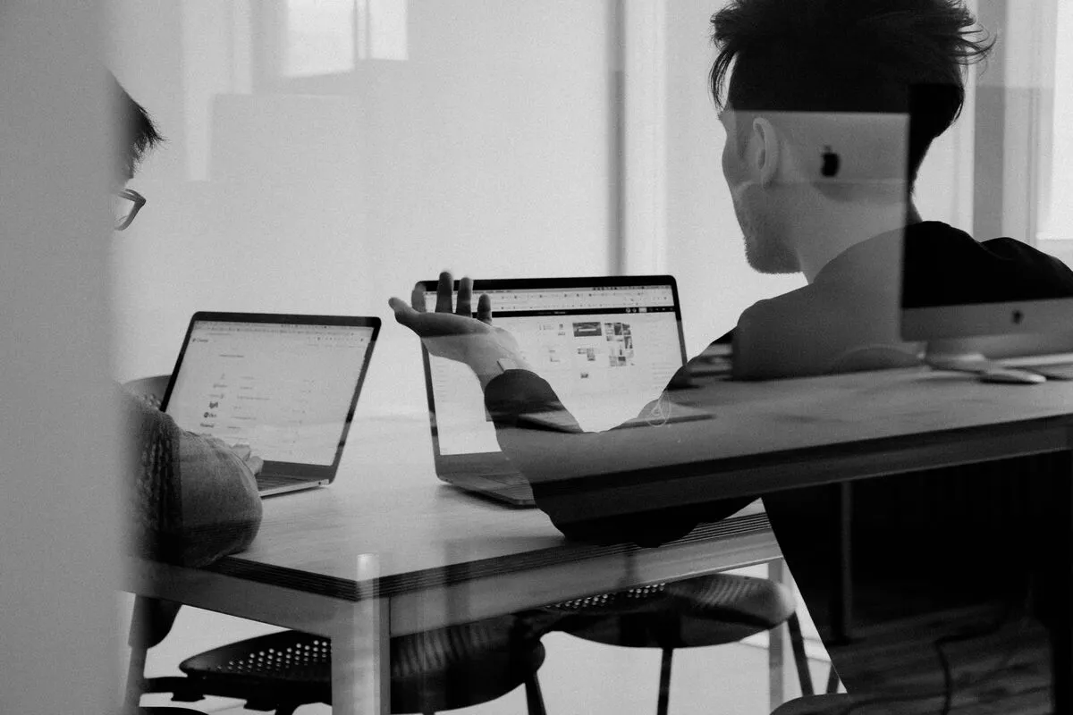 Two colleagues at laptops in discussion across a desk, one gesturing, in a black-and-white office scene.