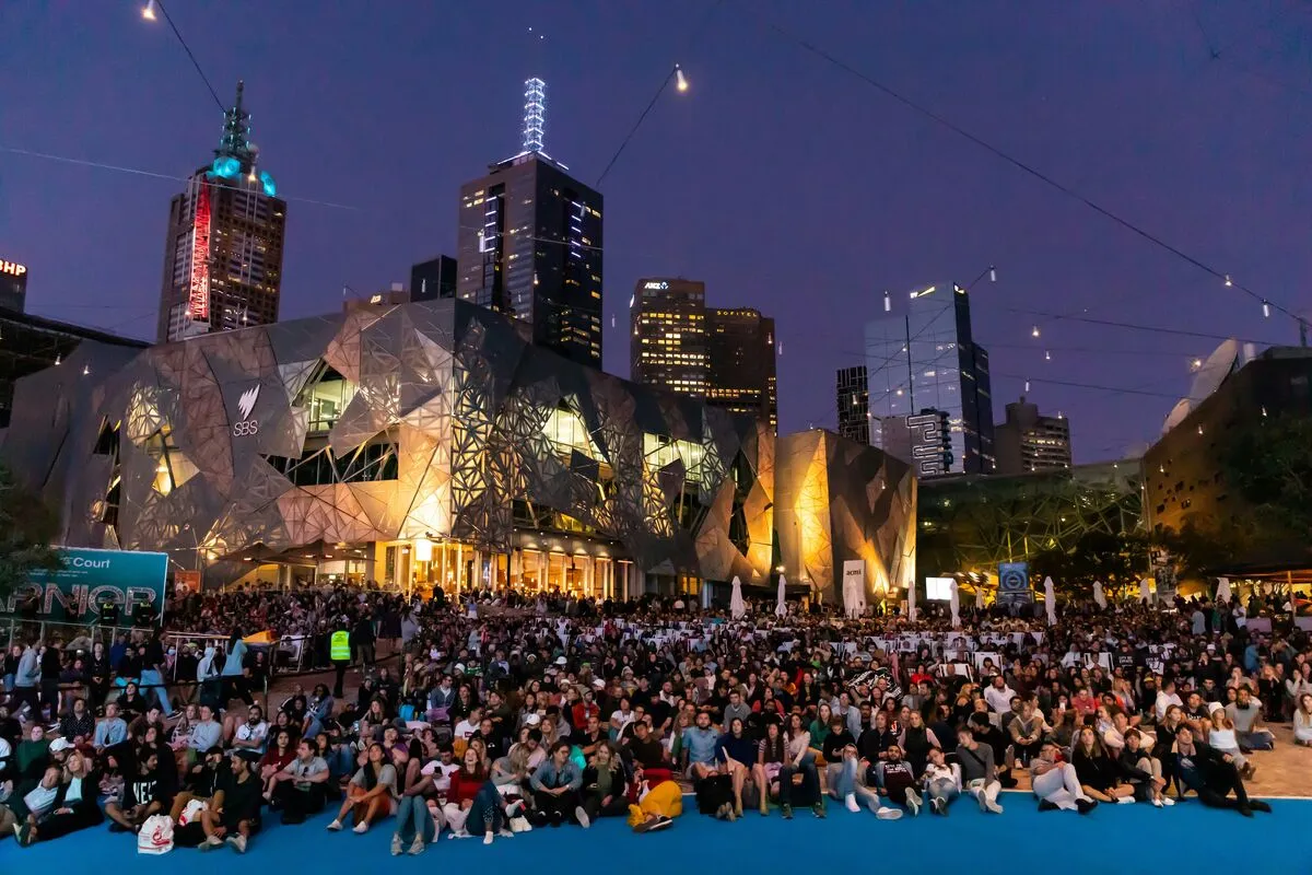 A large crowd gathered at Federation Square at dusk, with the lit Melbourne skyline behind them.
