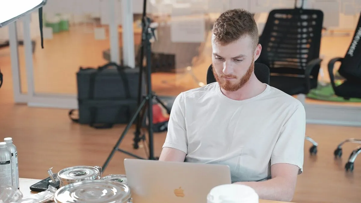 A man works intently on a laptop at a desk in a bright open-plan studio.