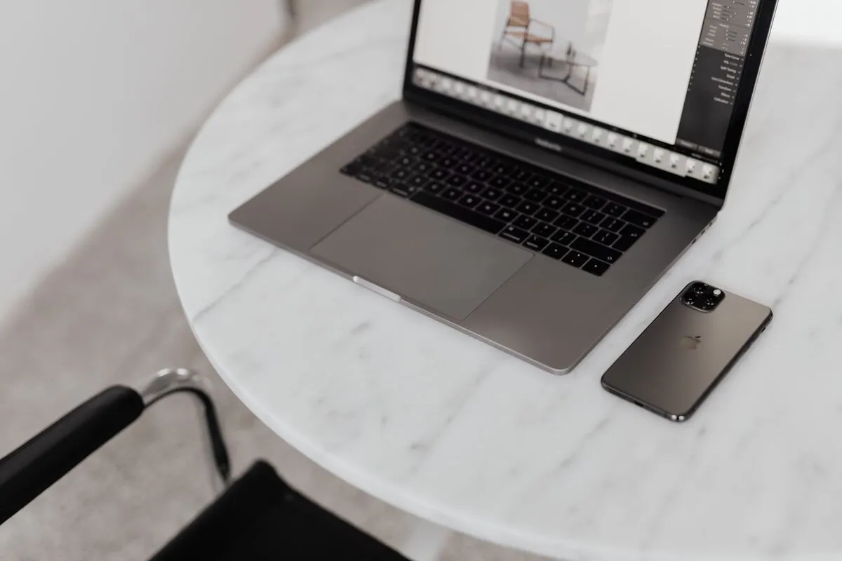 A laptop on a round marble table showing photo-editing software with a furniture product shot, a phone beside it.