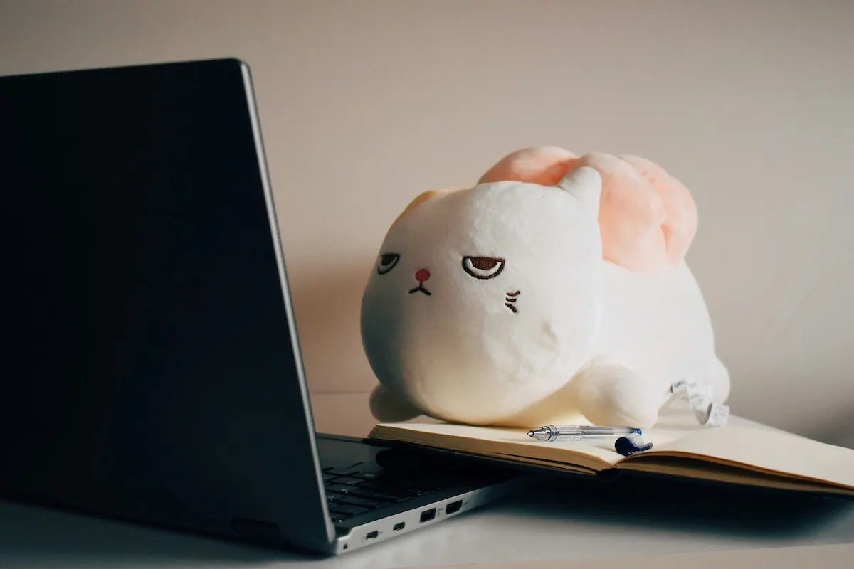 A grumpy-looking plush cat toy resting on an open notebook beside a laptop.