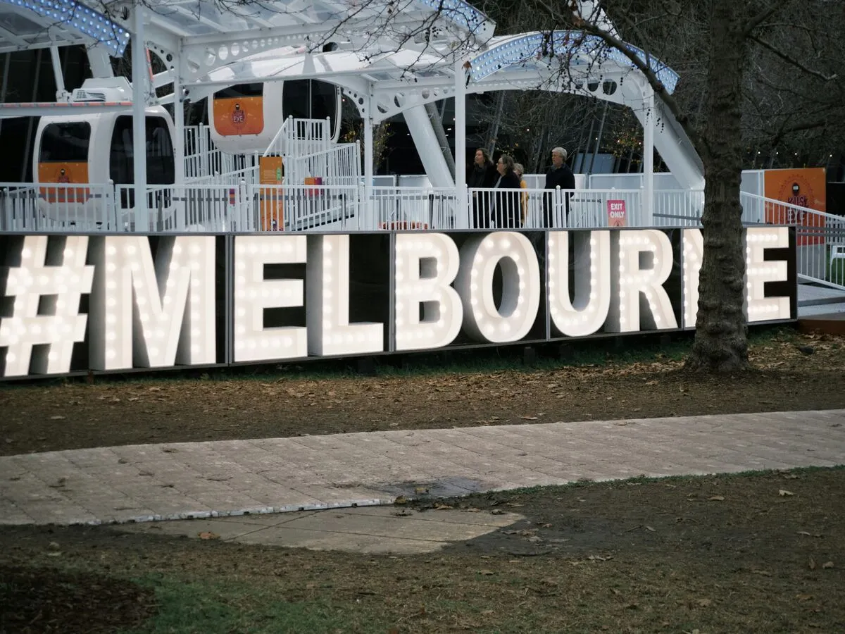 A large illuminated #MELBOURNE marquee sign in a park at dusk, with a Ferris wheel gondola behind it.