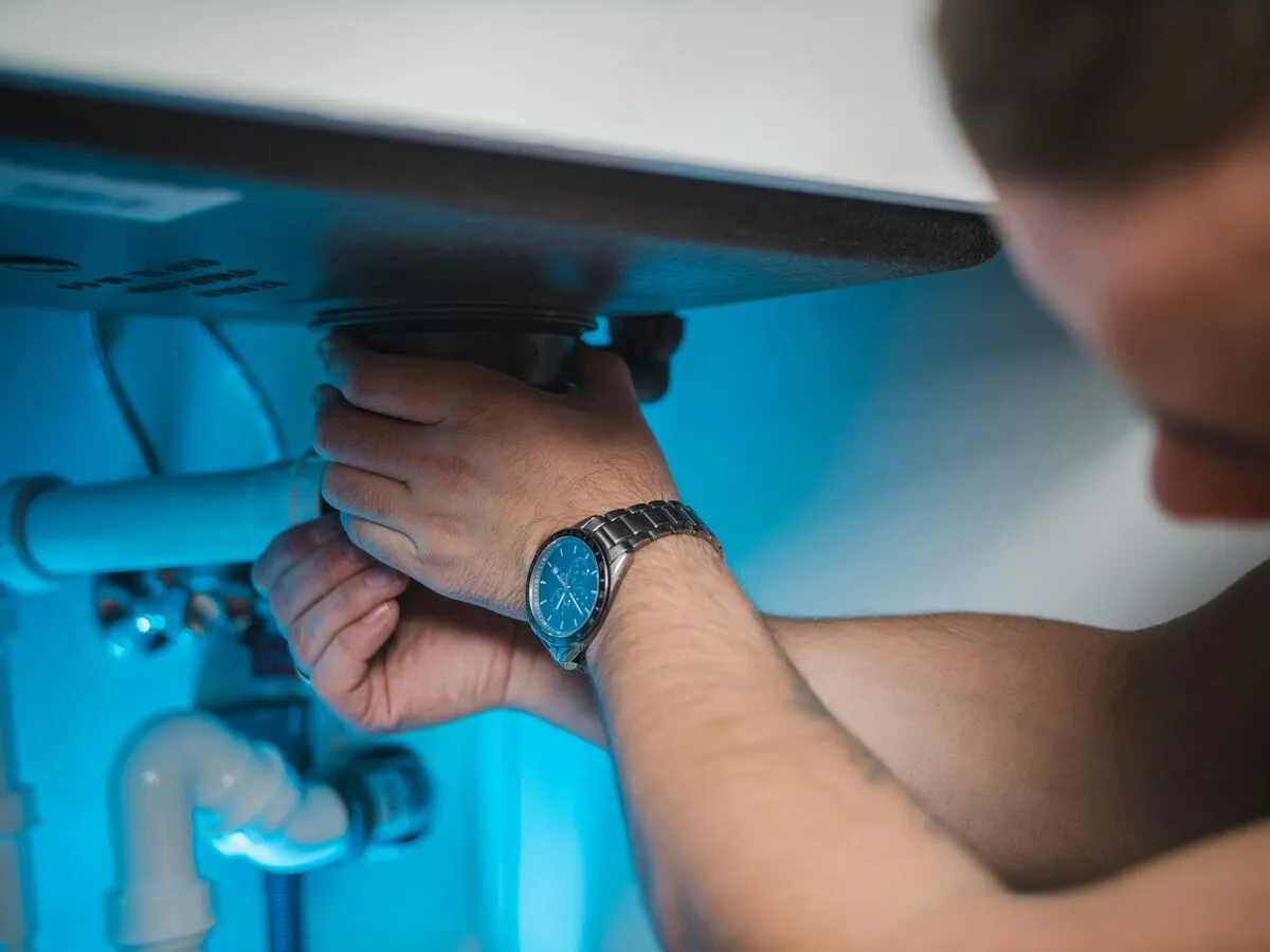 A plumber reaching under a sink to fix the pipework, wearing a steel wristwatch.