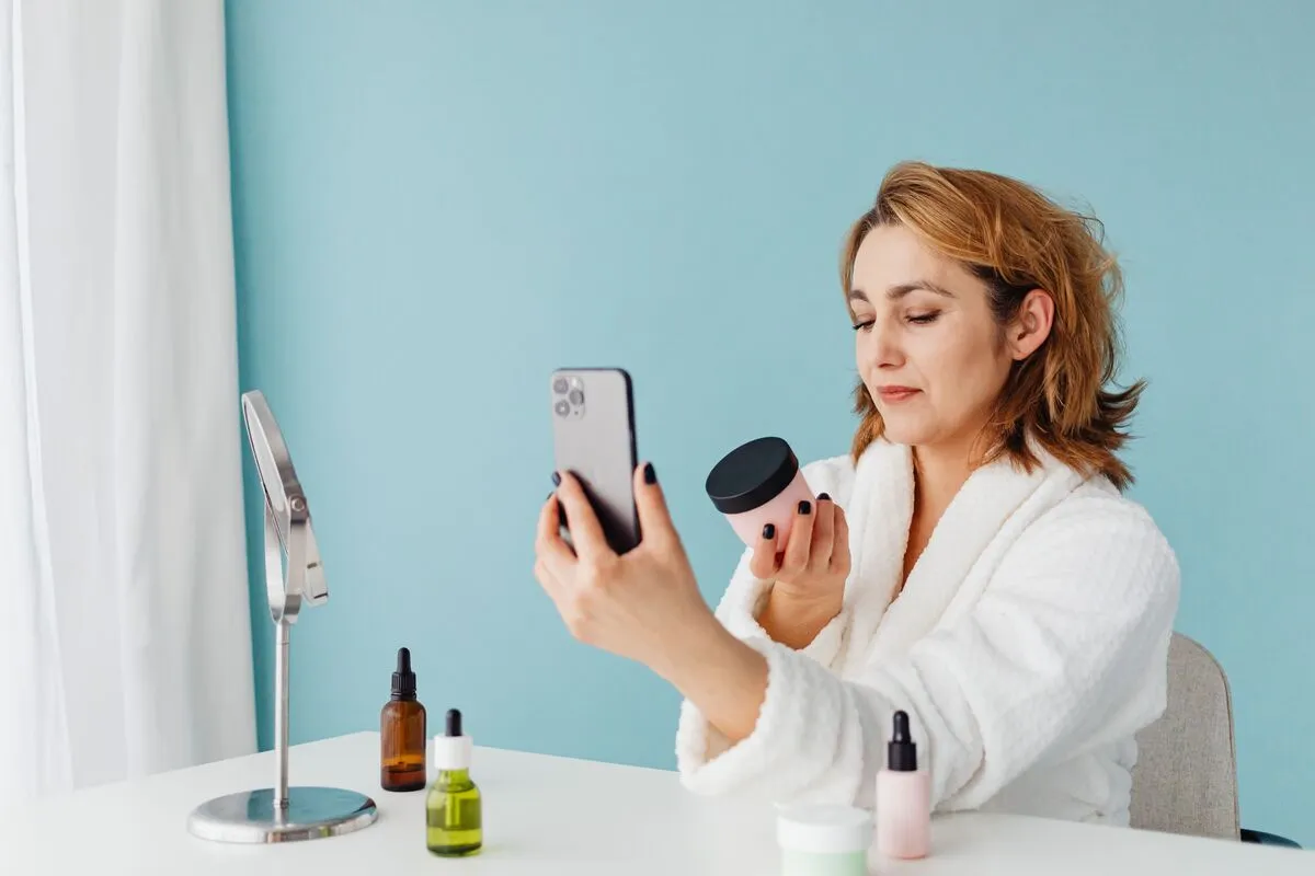 A woman in a bathrobe compares a jar of skincare against her phone at a dressing table, with serum bottles and a mirror in front of her.