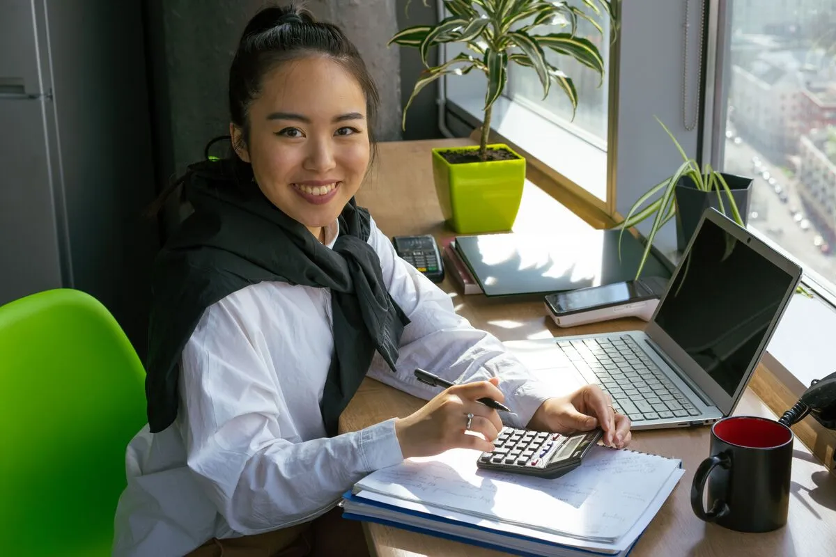 A small-business owner at a sunlit desk with a calculator, laptop, and paperwork.
