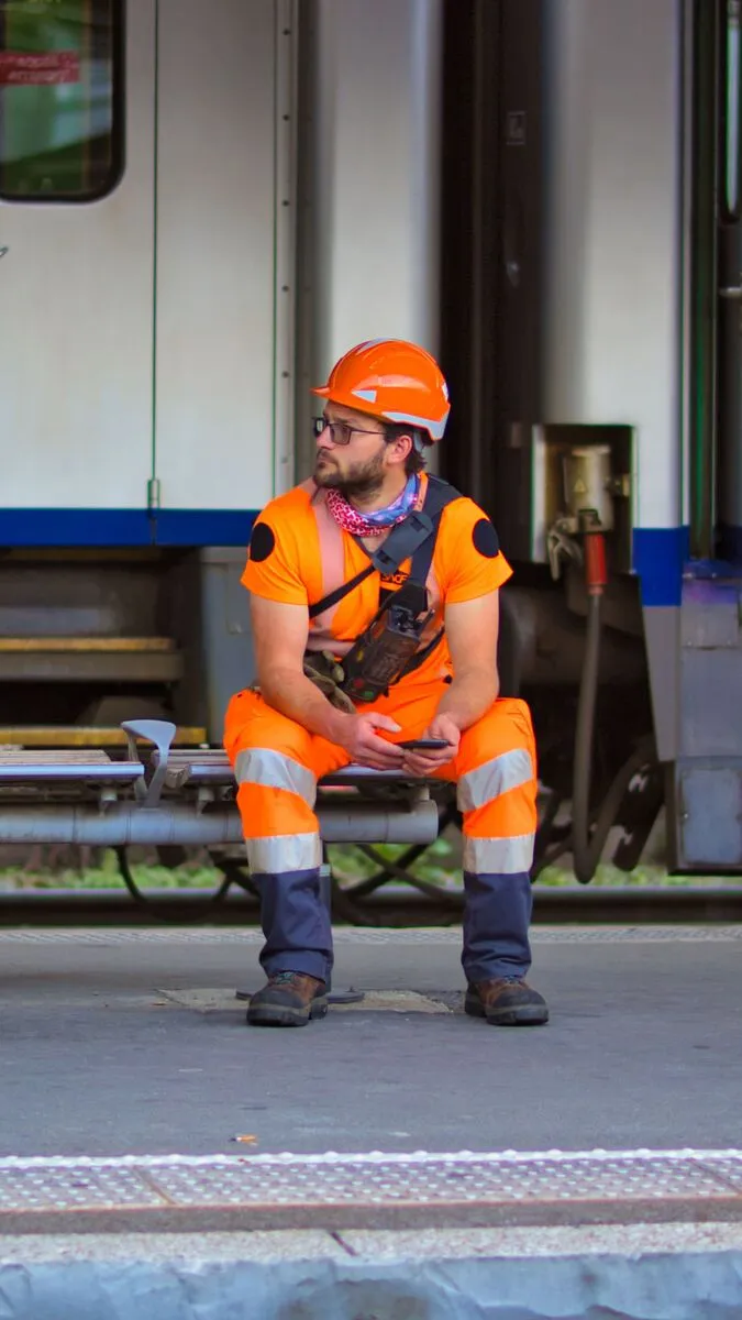 A worker in hi-vis orange sitting on a railway platform, checking their phone between jobs.