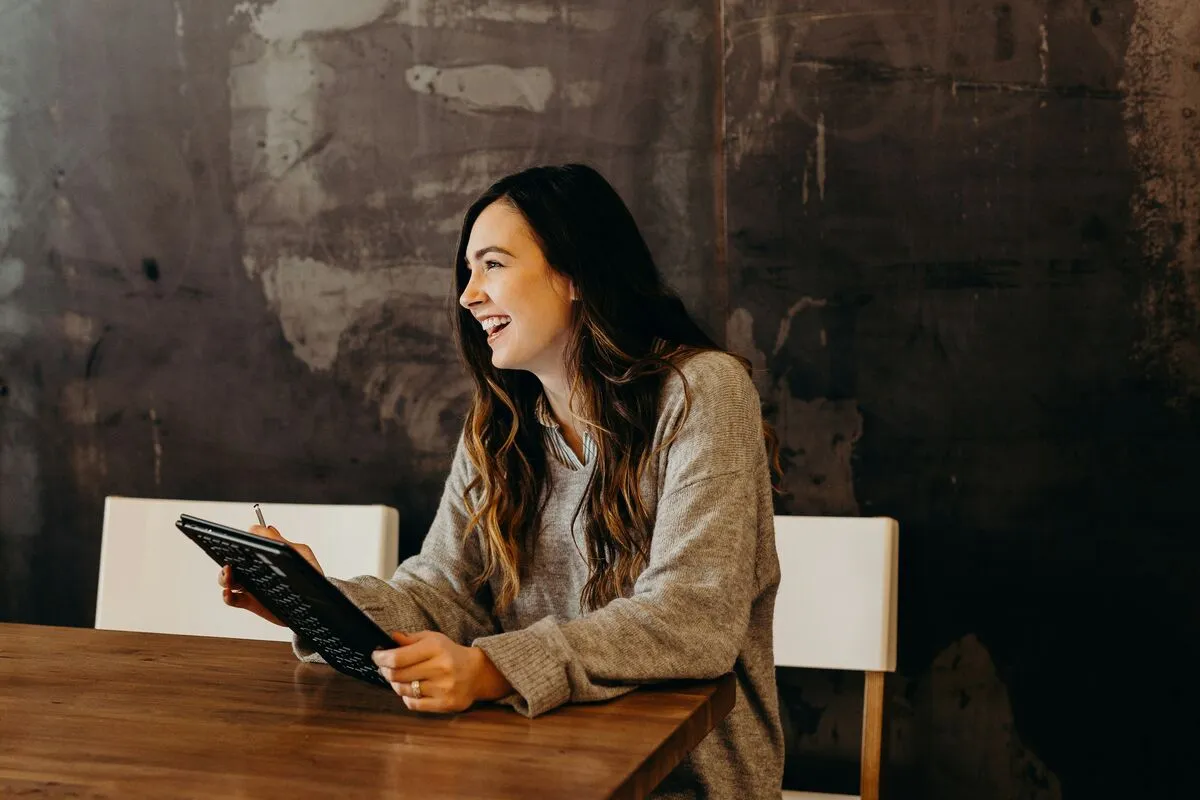 A smiling woman using a tablet with a stylus at a wooden table.