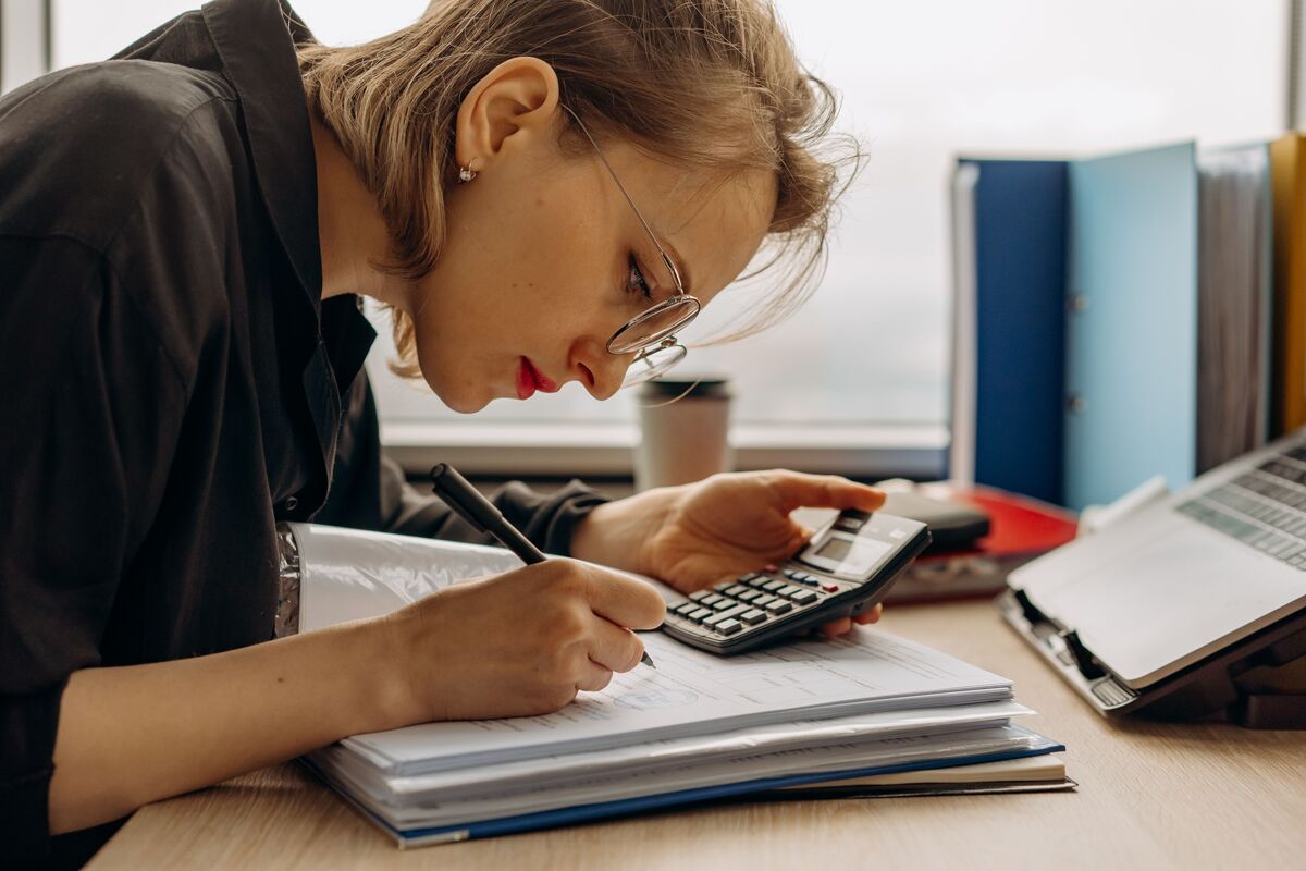 A woman in glasses doing the books with a calculator, a ledger, and folders.