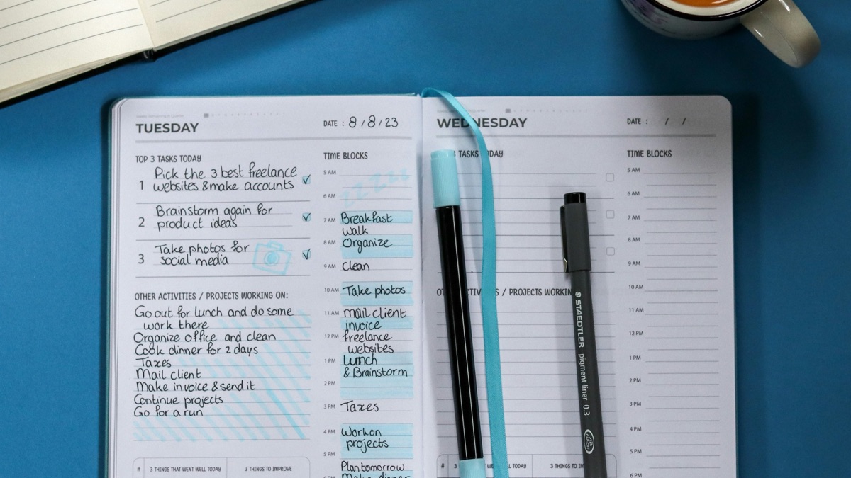 An open paper planner filled with handwritten daily tasks and time blocks, with pens and a coffee on the desk.