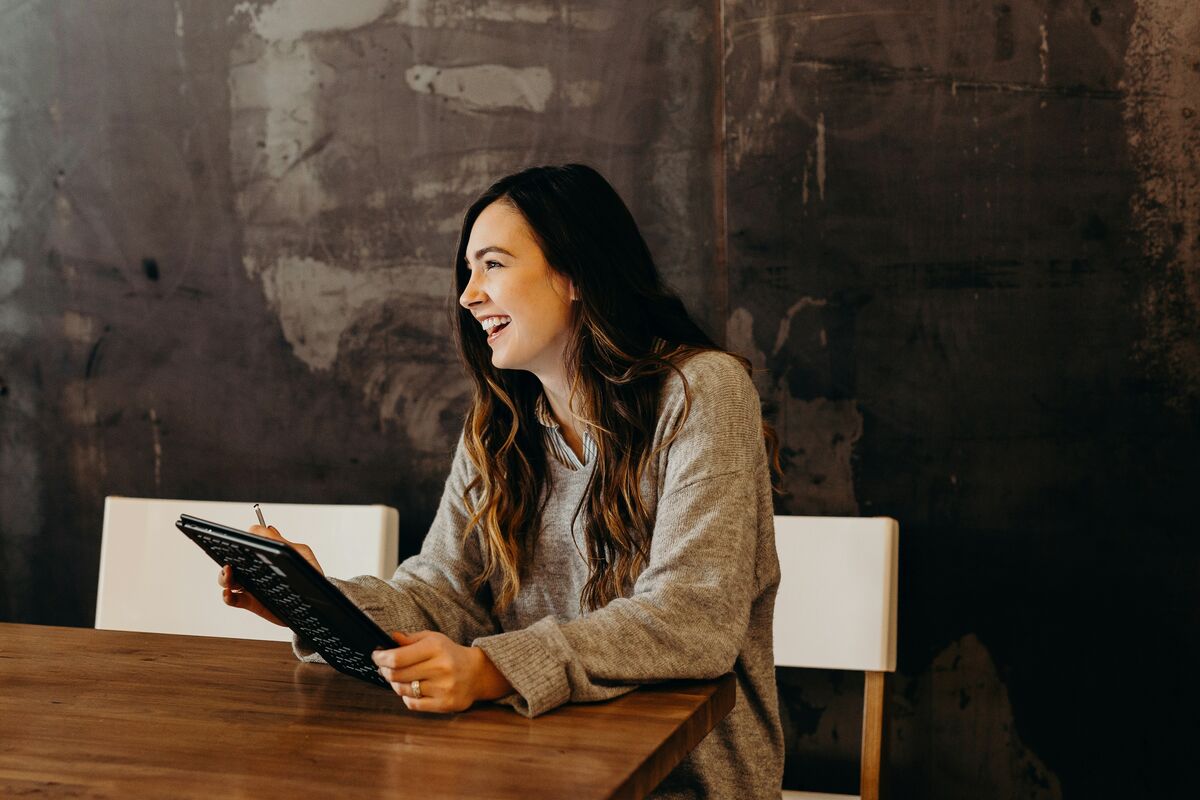 A smiling woman using a tablet with a stylus at a wooden table.